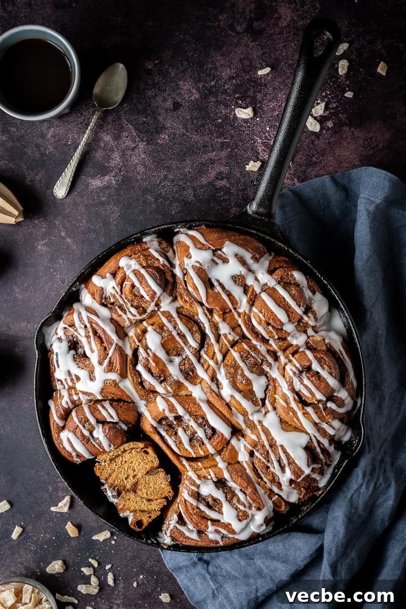 Soft vegan gingerbread cinnamon rolls in a skillet with ginger glaze a cast iron skillet of vegan gingerbread cinnamon rolls, one sliced in half, on a dark background with candied ginger and a grey cloth.