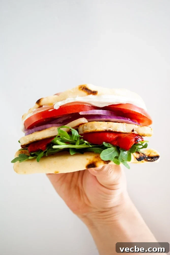 Grilled Halloumi Sandwich with Roasted Pepper and Spinach 2 A hand holding a sandwich of fried halloumi with roasted red pepper, arugula, tomato, red onion and tzatziki.