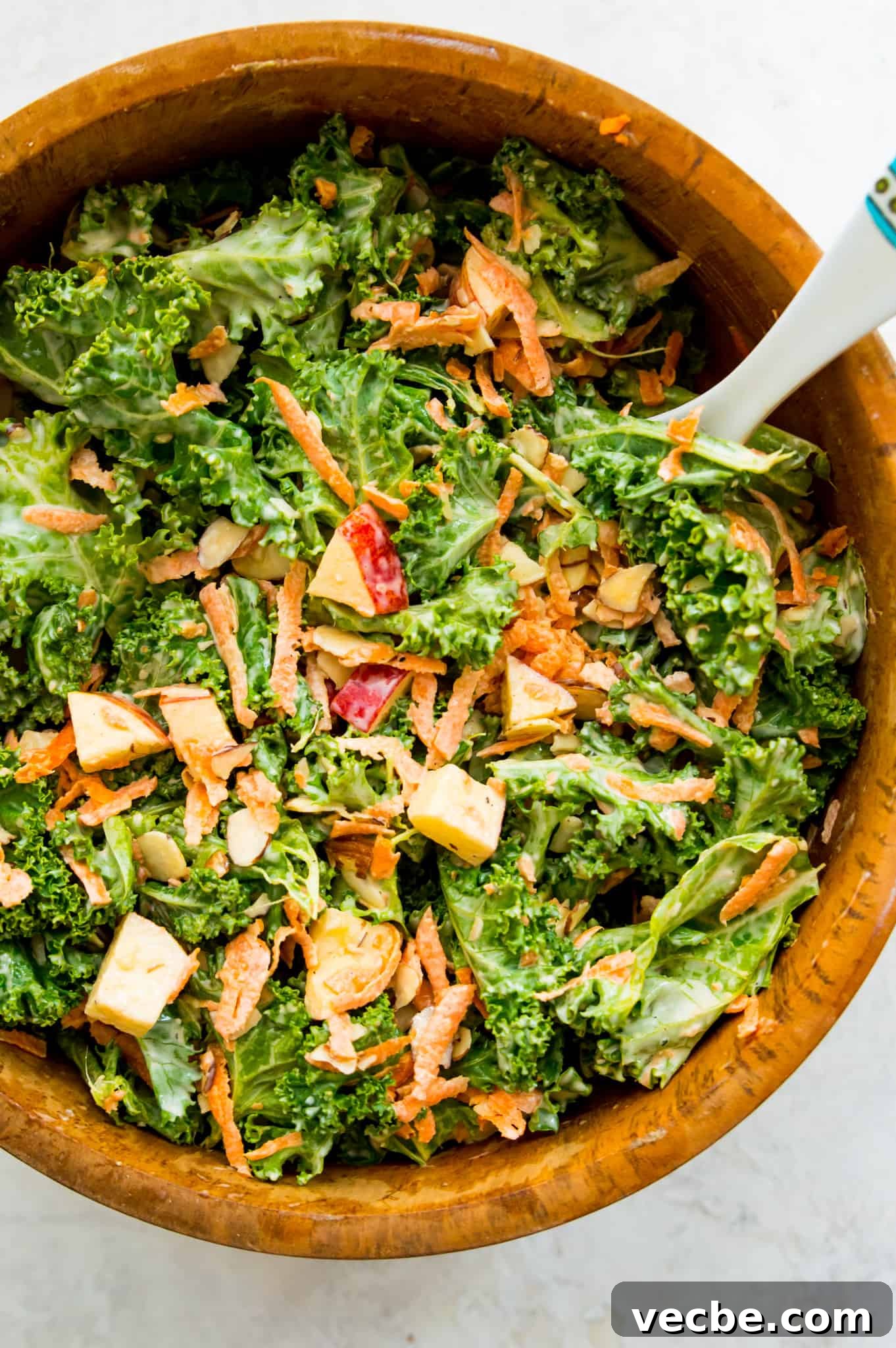 Kale Slaw with Apples Pieces of red apple, carrots, slivered almonds, and fresh kale in a wooden mixing bowl with dressing.