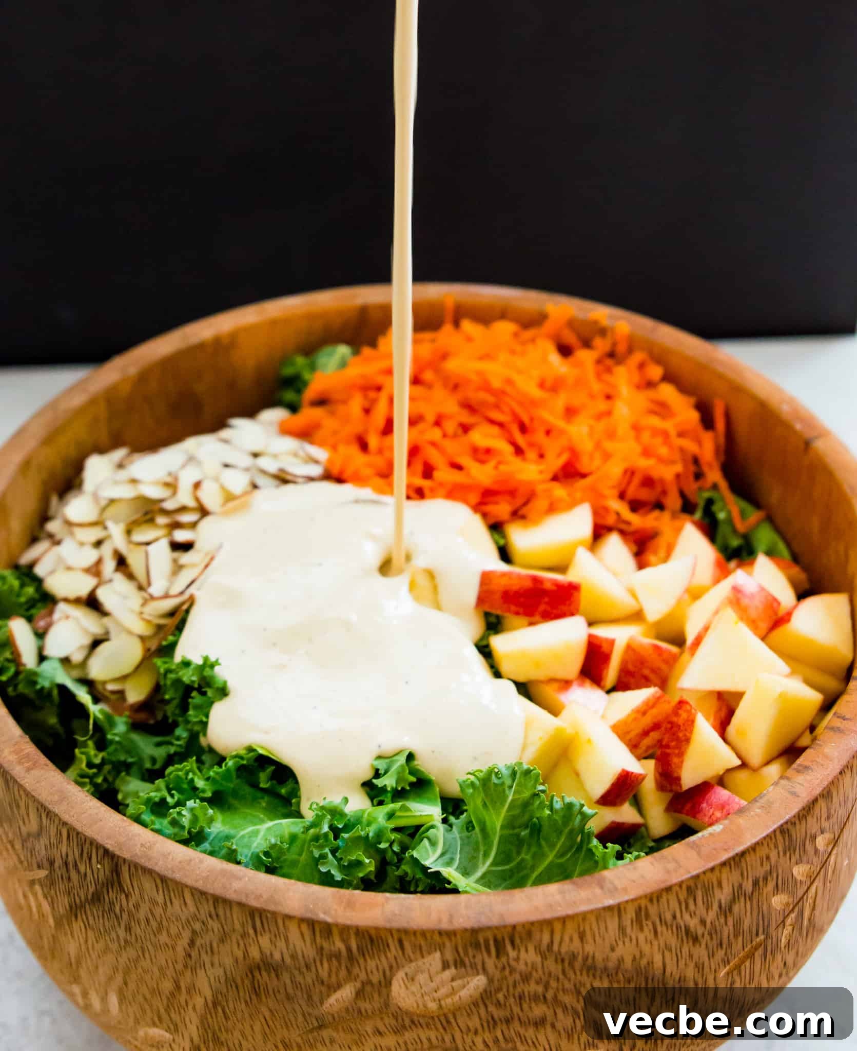 Cabbage-Free Kale Slaw with Apples and Carrots 6 A kale salad in a wooden bowl with a creamy white dressing being poured on it.
