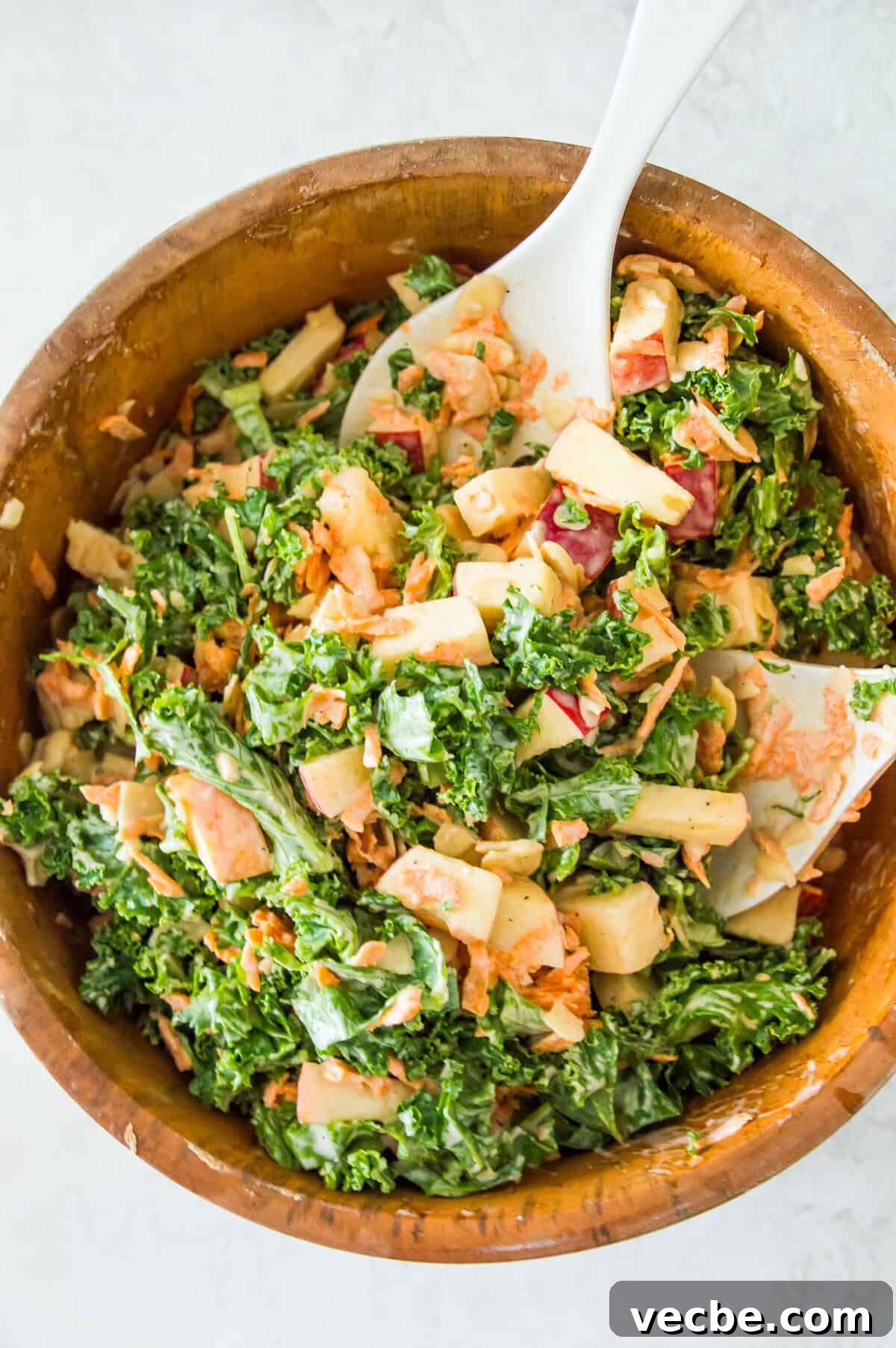 Cabbage-Free Kale Slaw with Apples and Carrots 7 A wooden salad bowl with a kale and apple salad in it and two serving spoons in the bowl.