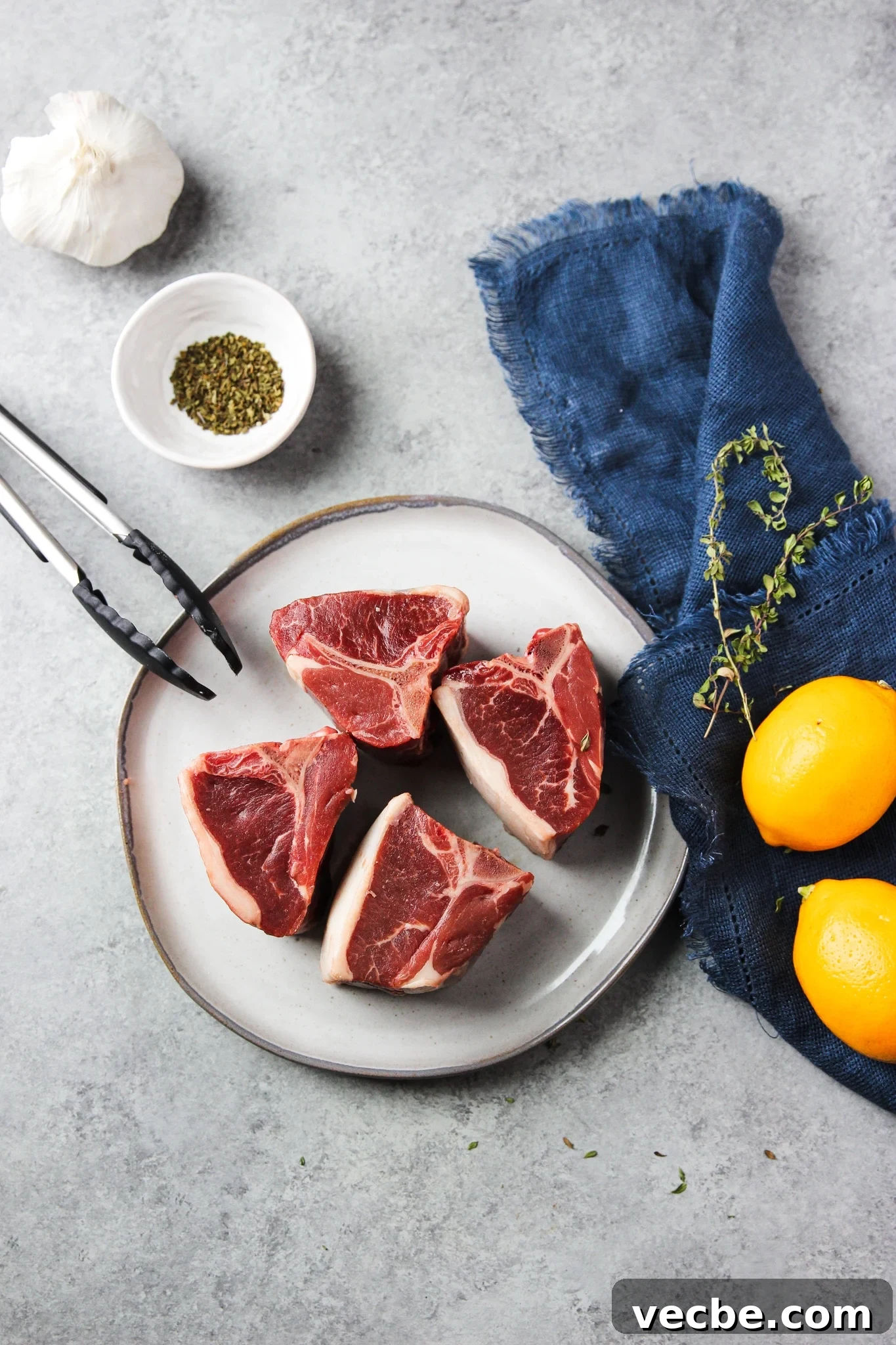 Pan-Seared Garlic & Herb Lamb Loin Chops 3 4 raw lamb loin chops on a white plate next to lemon, oregano, and small white bowls of herbs.