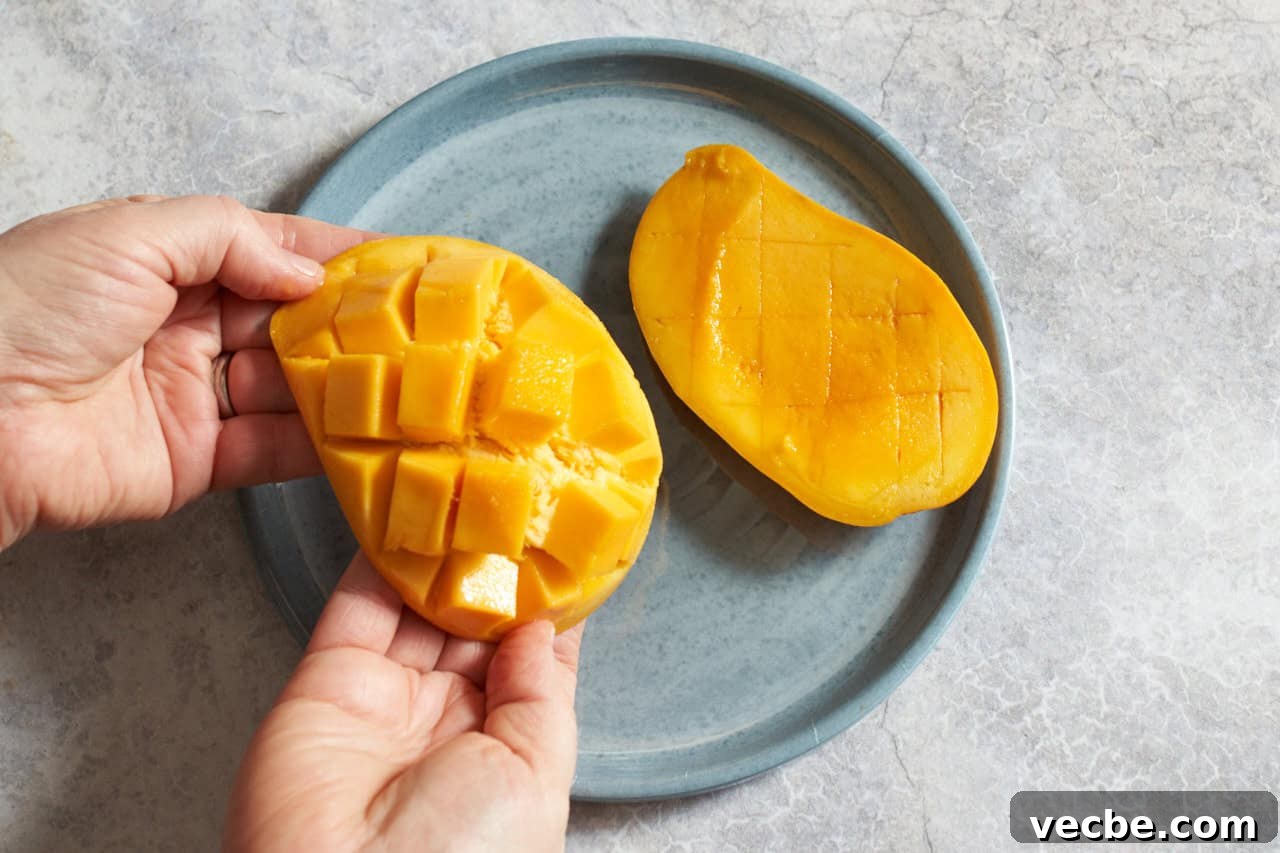 How to Cut a Mango Like a Pro 2 A woman's hands are shown bending a mango that has been scored so that cube-shaped pieces of the fruit are releasing from the peel, another mango half is on a plate to the right.
