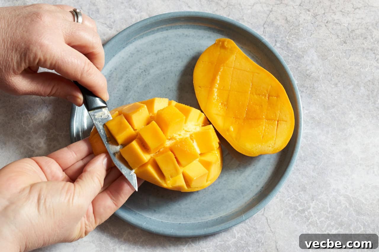 How to Cut a Mango Like a Pro 3 A woman's hands are shown slicing cube-shaped pieces of mango away from its peel, another mango half is on a plate to the right.