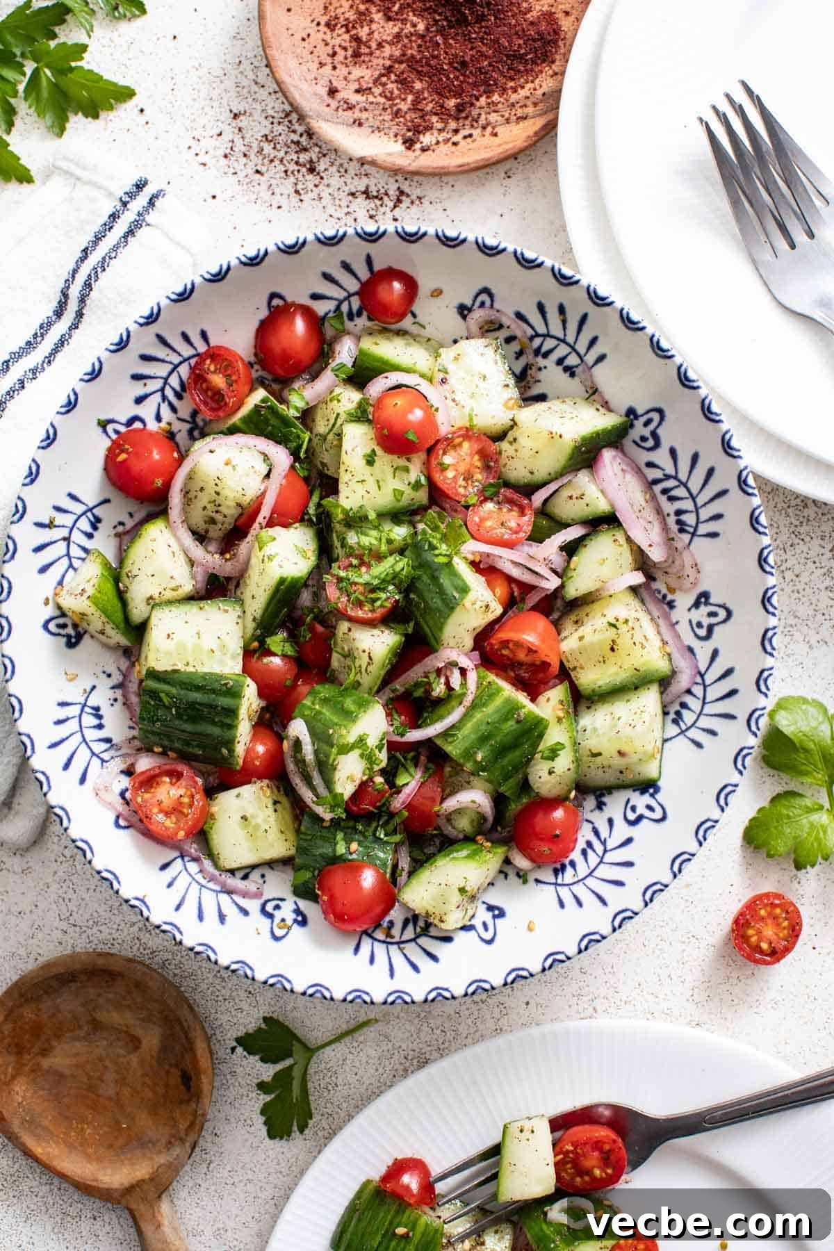 Refreshing Mediterranean Cucumber Salad with Feta and Mint 2 Cucumber and tomato salad in a white and blue bowl next to plates, a wood spoon, and a bowl of sumac.