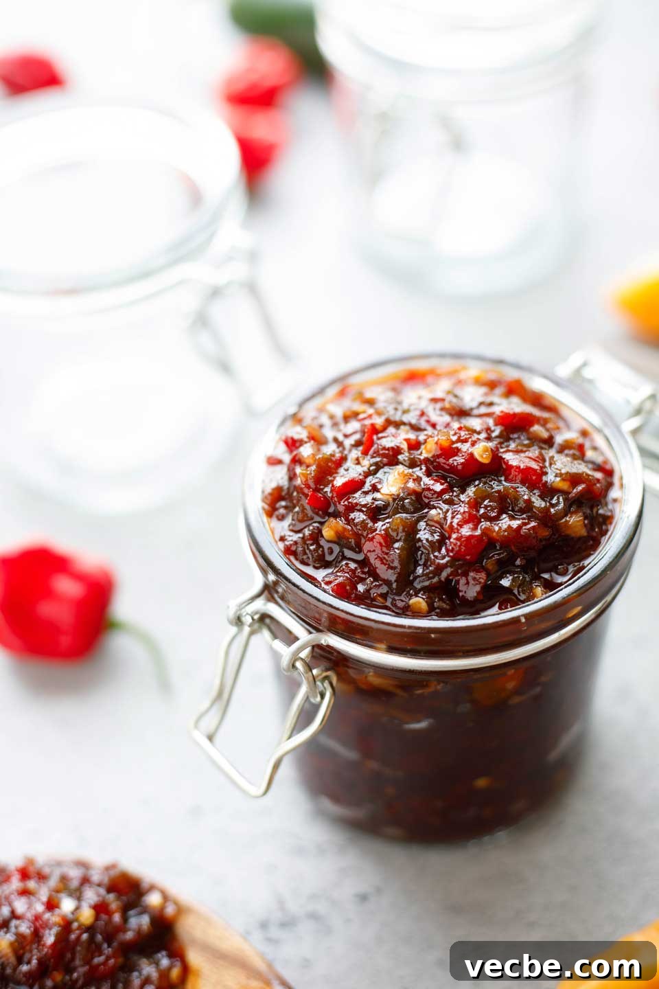 Homemade Sweet and Spicy Pepper Relish Recipe 2 Glass jar full of relish with empty jars and raw, hot peppers in background.