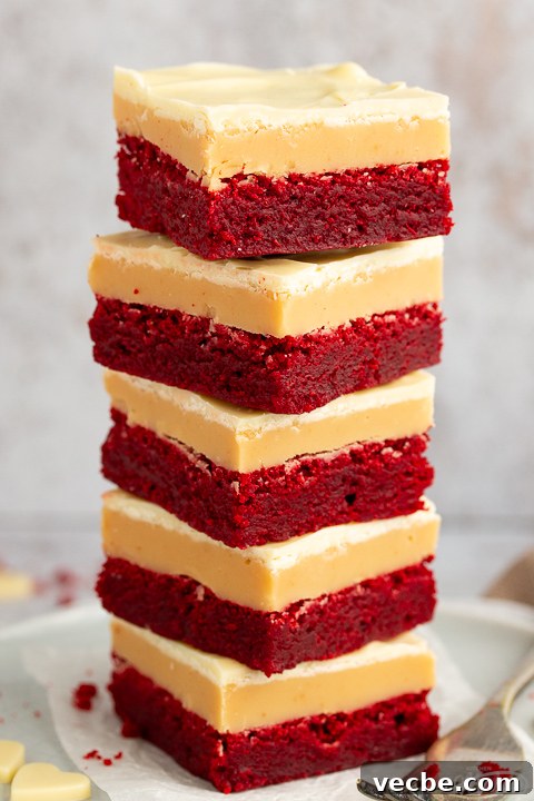 Red Velvet Millionaire Brownies Recipe with Caramel and Chocolate 2 A stack of 5 red velvet millionaire brownies set on two stacked plates with a fork on the side. There are white chocolate hearts, red velvet brownie crumbs and a light brown napkin in the background.