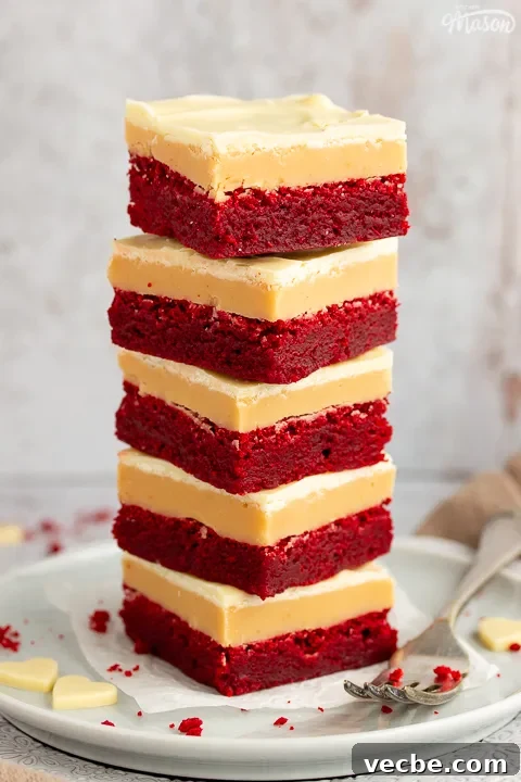 Red Velvet Millionaire Brownies Recipe with Caramel and Chocolate 5 A stack of 5 red velvet millionaire brownies set on two stacked plates with a fork on the side. There are white chocolate hearts, red velvet brownie crumbs and a light brown napkin in the background.