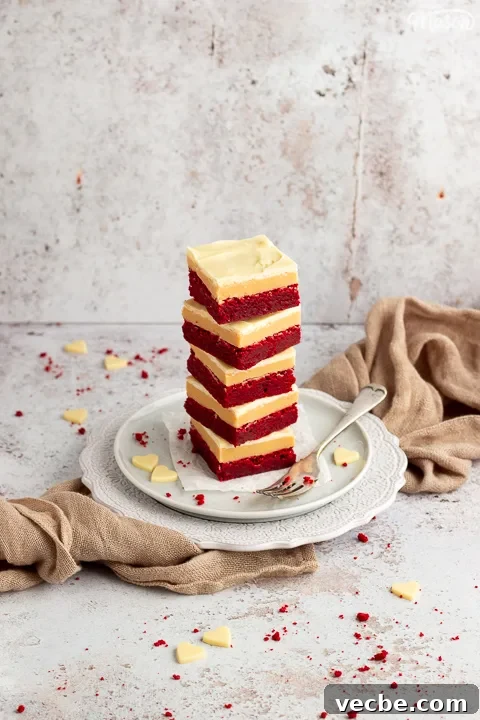 Red Velvet Millionaire Brownies Recipe with Caramel and Chocolate 6 A stack of 5 red velvet millionaire brownies set on two stacked plates with a fork on the side. There are white chocolate hearts, red velvet brownie crumbs and a light brown napkin in the background.