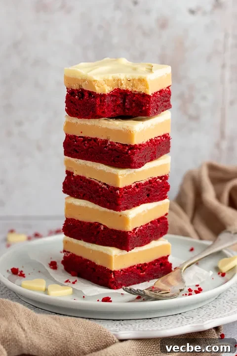 Red Velvet Millionaire Brownies Recipe with Caramel and Chocolate 8 A stack of 5 red velvet millionaire brownies set on two stacked plates with a fork on the side. There are white chocolate hearts, red velvet brownie crumbs and a light brown napkin in the background.