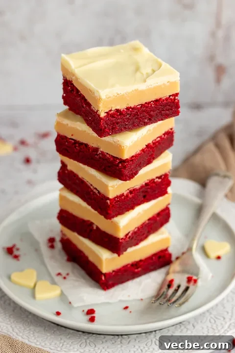 Red Velvet Millionaire Brownies Recipe with Caramel and Chocolate 9 A stack of 5 red velvet millionaire brownies set on two stacked plates with a fork on the side. There are white chocolate hearts, red velvet brownie crumbs and a light brown napkin in the background.