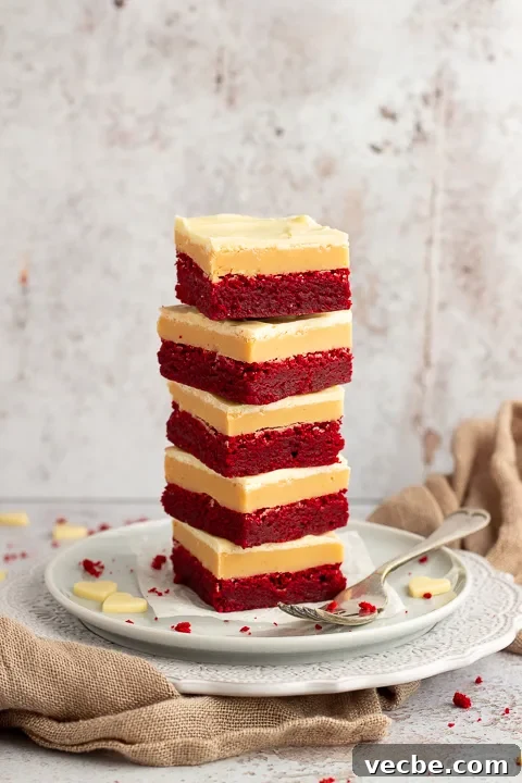 Red Velvet Millionaire Brownies Recipe with Caramel and Chocolate 4 A stack of 5 red velvet millionaire brownies set on two stacked plates with a fork on the side. There are white chocolate hearts, red velvet brownie crumbs and a light brown napkin in the background.