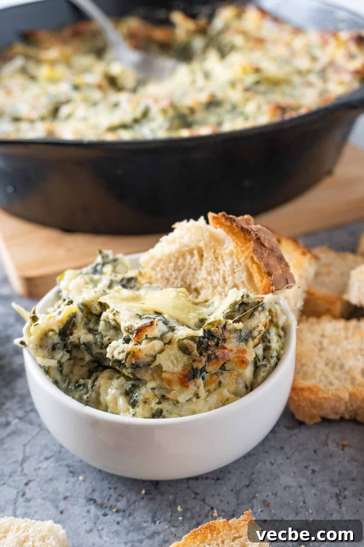 Creamy Hot Artichoke Dip Recipe 2 Hot artichoke Dip in a small white bowl with a piece of bread sticking in it.