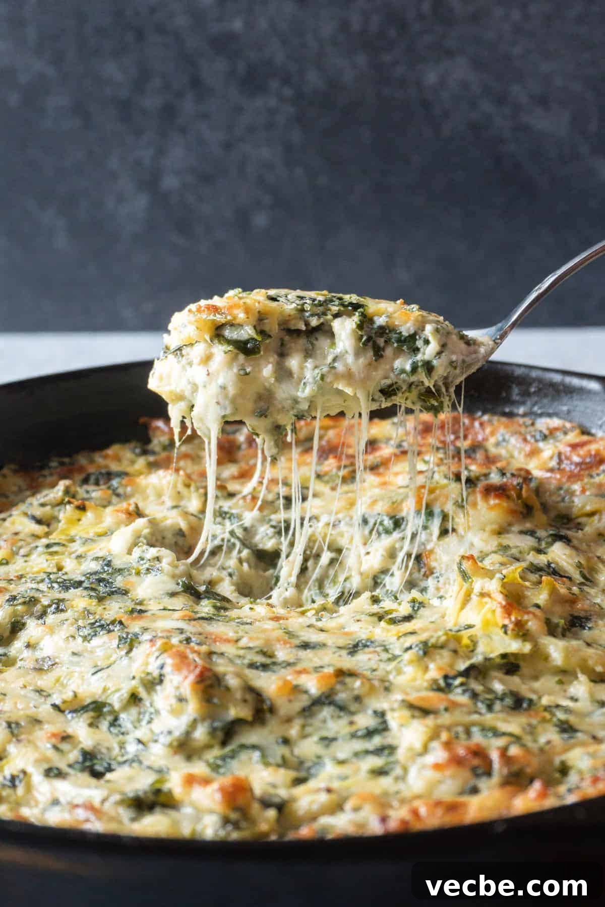 Creamy Hot Artichoke Dip Recipe 4 Spoon full of Hot Artichoke Dip with a a cheesy pull connecting the spoon to the dip in a cast iron pan.