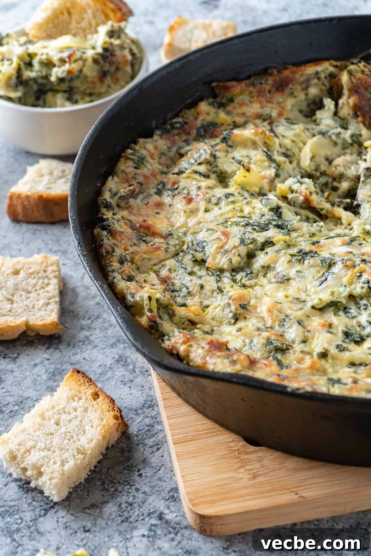 Creamy Hot Artichoke Dip Recipe 10 Hot artichoke dip in a cast iron pan on wooden trivet with pieces of bread scattered around.