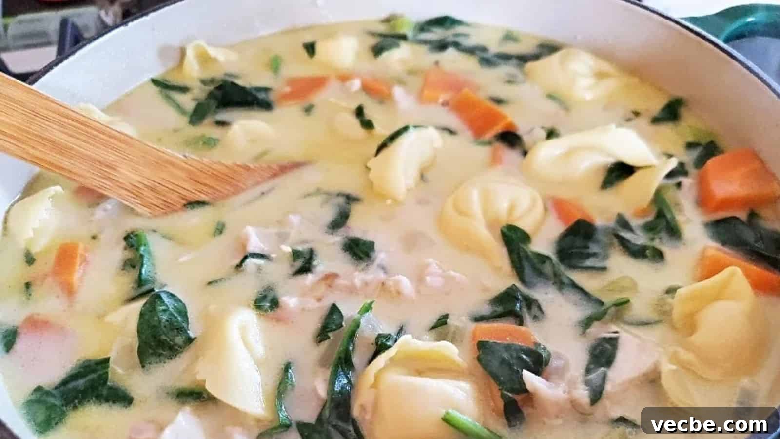 19 Budget-Friendly Meals to Replace Takeout 20 Image shows Chicken tortellini soup in a ceramic enameled pot with a wooden spoon stirring it.