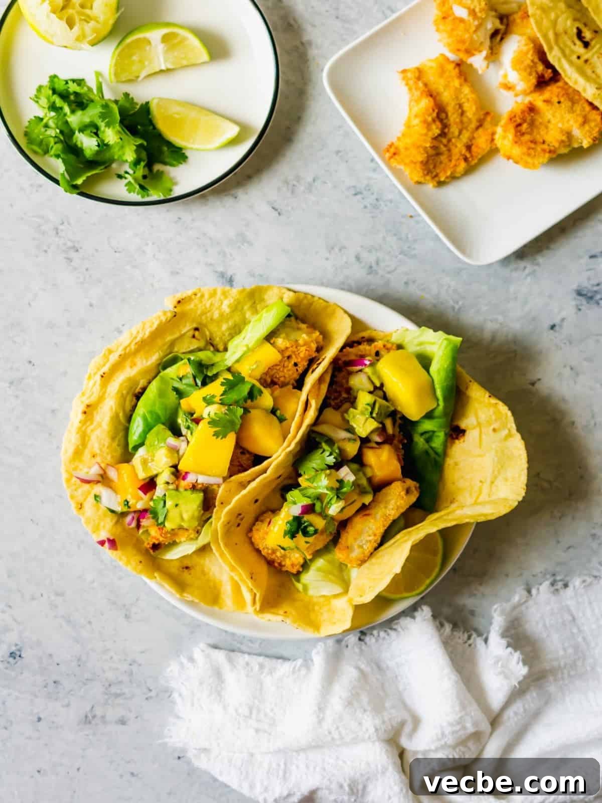 Crispy Fish Tacos with Fresh Mango Salsa Recipe 2 Two fish tacos topped with mango salsa next to a plate of lime wedges and cilantro.