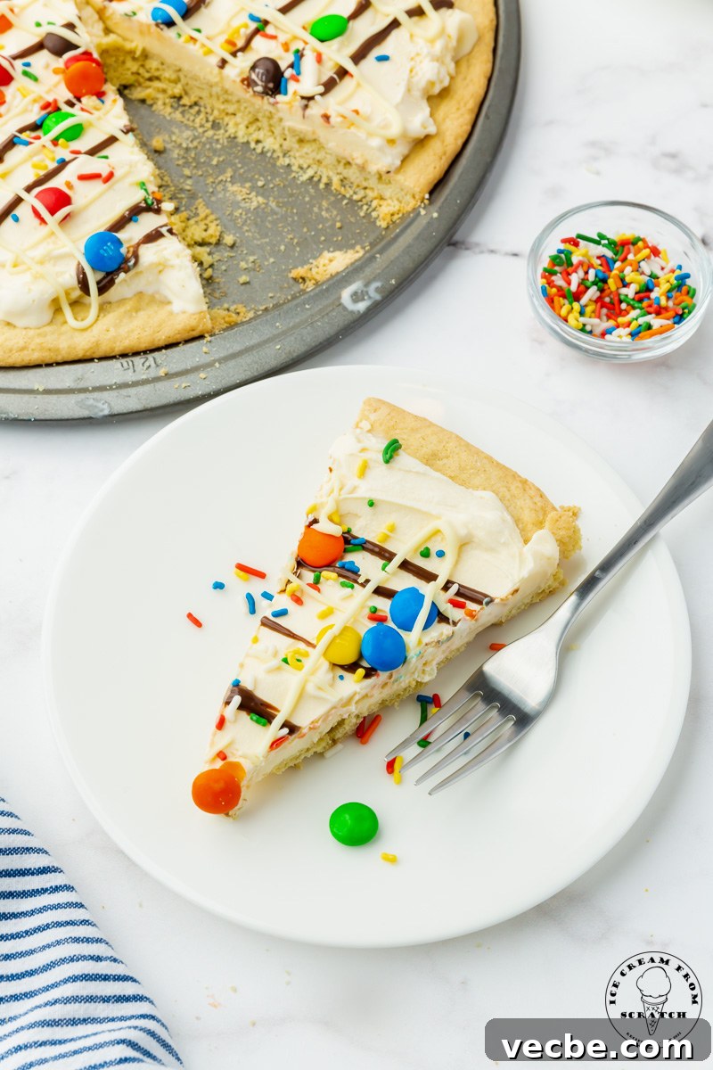 No-Bake Ice Cream Pizza Recipe with Cookie Crust 6 a white plate holding a slice of ice cream pizza and a fork. behind the plate is a small bowl of rainbow sprinkles and the remaining cookie ice cream pizza on a pizza pan.
