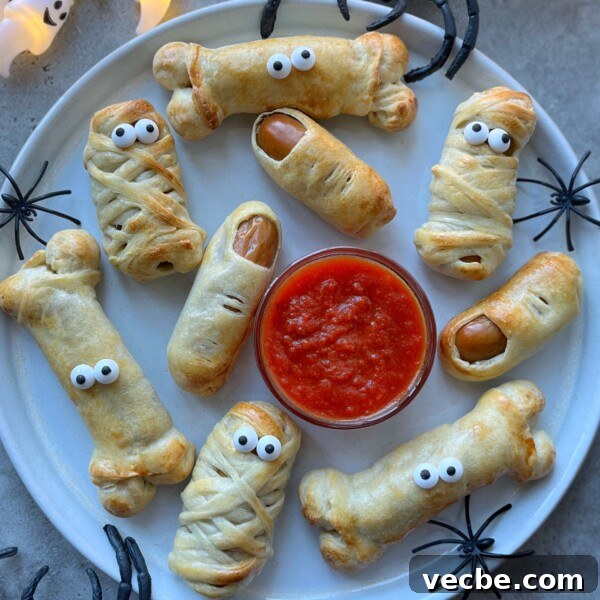 Spooky Halloween Appetizers to Wow Party Guests 4 A plate of Halloween Sausage Rolls shaped like mummies and bones, made from pastry-wrapped sausages with candy eyes, surrounds a bowl of red dipping sauce. Plastic spiders are placed around the plate for a spooky touch.