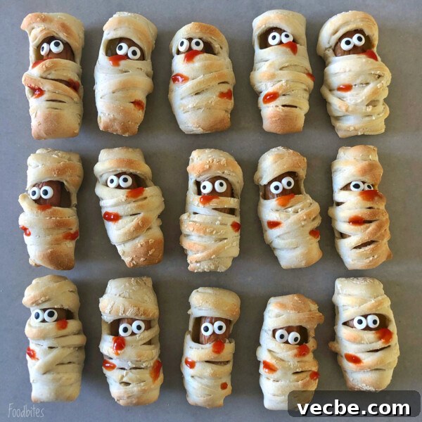 Spooky Halloween Appetizers to Wow Party Guests 7 Rows of mummy-shaped bites with googly candy eyes and streaks of ketchup for mouths, arranged neatly on a grey surface. Each pastry is wrapped in dough to resemble a mummy.