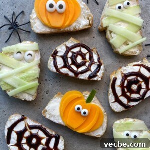 Spooky Halloween Appetizers to Wow Party Guests 8 Slices of bread with cream cheese are decorated for Halloween as mummies, spiders, and pumpkins using cucumber, yellow vegetables, balsamic glaze, and candy eyes. Plastic spiders are placed around the snacks.