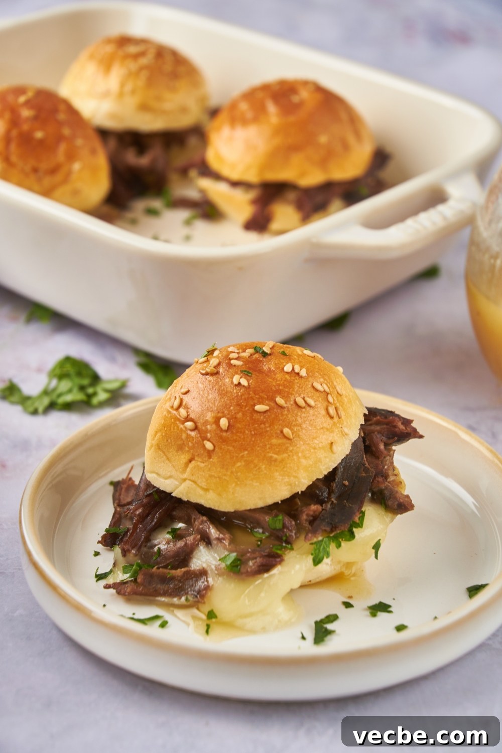 Fall-Apart Slow Cooker Tri-Tip Recipe 2 A mini tri tip sandwich filled with shredded beef, melted cheese, fresh green herbs, and topped with a poppyseed bun. In the background are three more tri tip sandwiches.