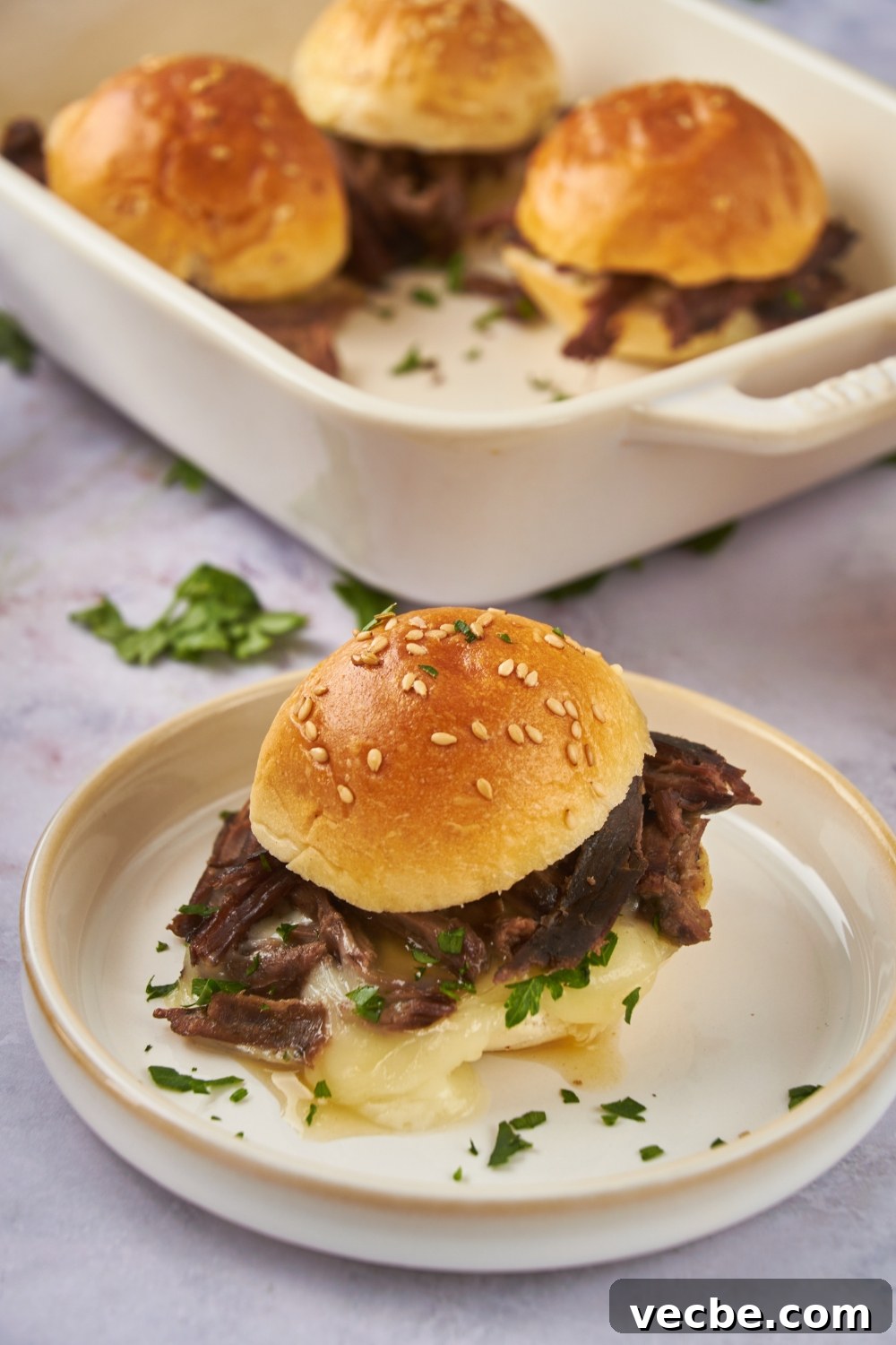 Fall-Apart Slow Cooker Tri-Tip Recipe 11 A mini tri tip sandwich filled with shredded beef, melted cheese, fresh green herbs, and topped with a poppyseed bun. In the background are three more tri tip sandwiches.