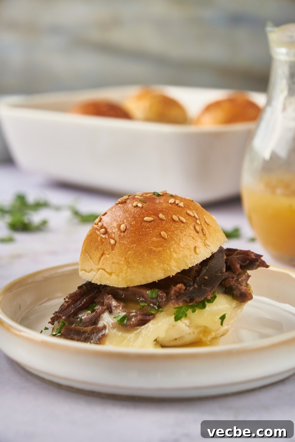 Fall-Apart Slow Cooker Tri-Tip Recipe 3 A mini tri tip sandwich filled with shredded beef, melted cheese, fresh green herbs, and topped with a poppyseed bun. In the background are three more tri tip sandwiches.