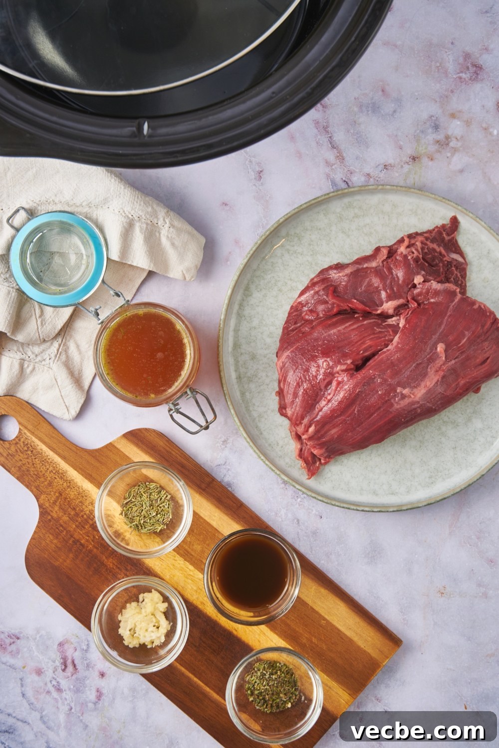 Fall-Apart Slow Cooker Tri-Tip Recipe 5 Overhead view of an assortment of ingredients including a raw tri tip roast and bowls of broth, garlic, Worcestershire sauce, and spices.