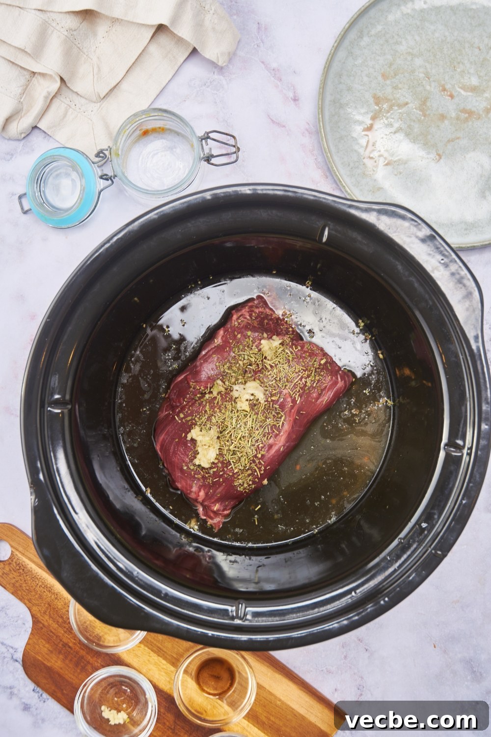 Fall-Apart Slow Cooker Tri-Tip Recipe 6 A raw beef roast in a black crock pot topped with minced garlic, dried herbs, and broth poured around the beef.