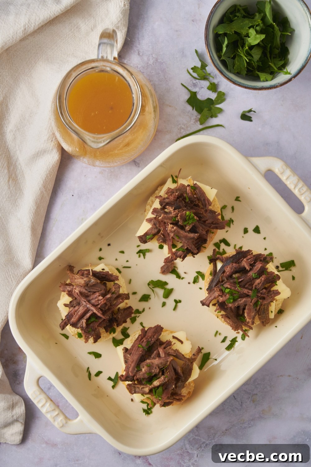 Fall-Apart Slow Cooker Tri-Tip Recipe 9 Overhead view of a white baking dish and four tri tip sandwiches layered with buns, cheese, tri tip, and fresh herbs.