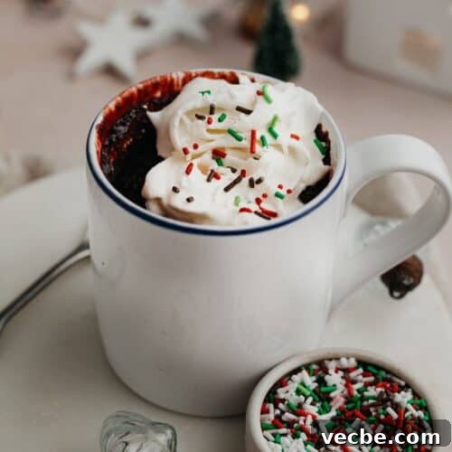 A red velvet mug cake topped with whipped cream and red and green sprinkles.