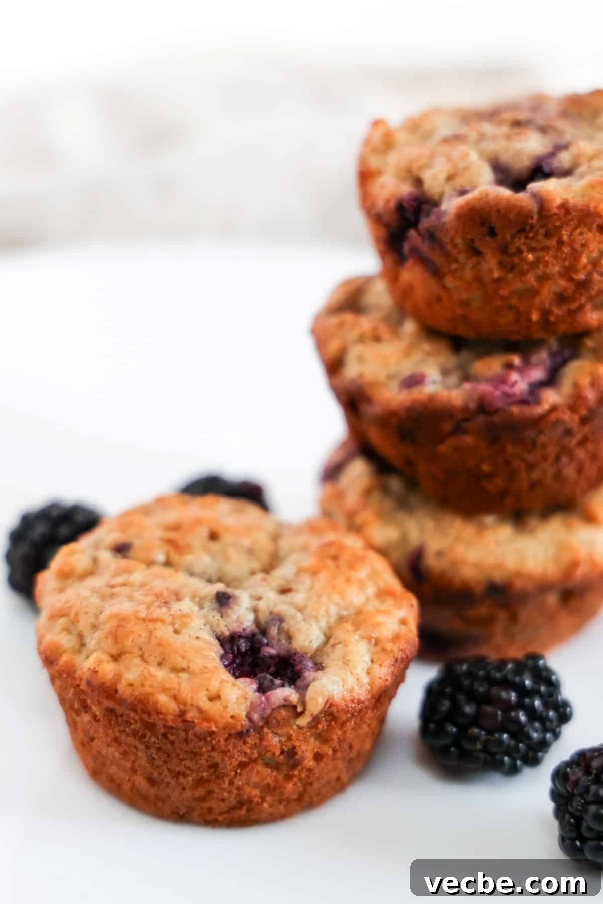 Banana Blackberry Oatmeal Muffins Recipe 2 Four blackberry banana oatmeal muffins, three are stacked on top of eachother to the right and one muffin is in the center. A few blackberries are scattered around the muffins.