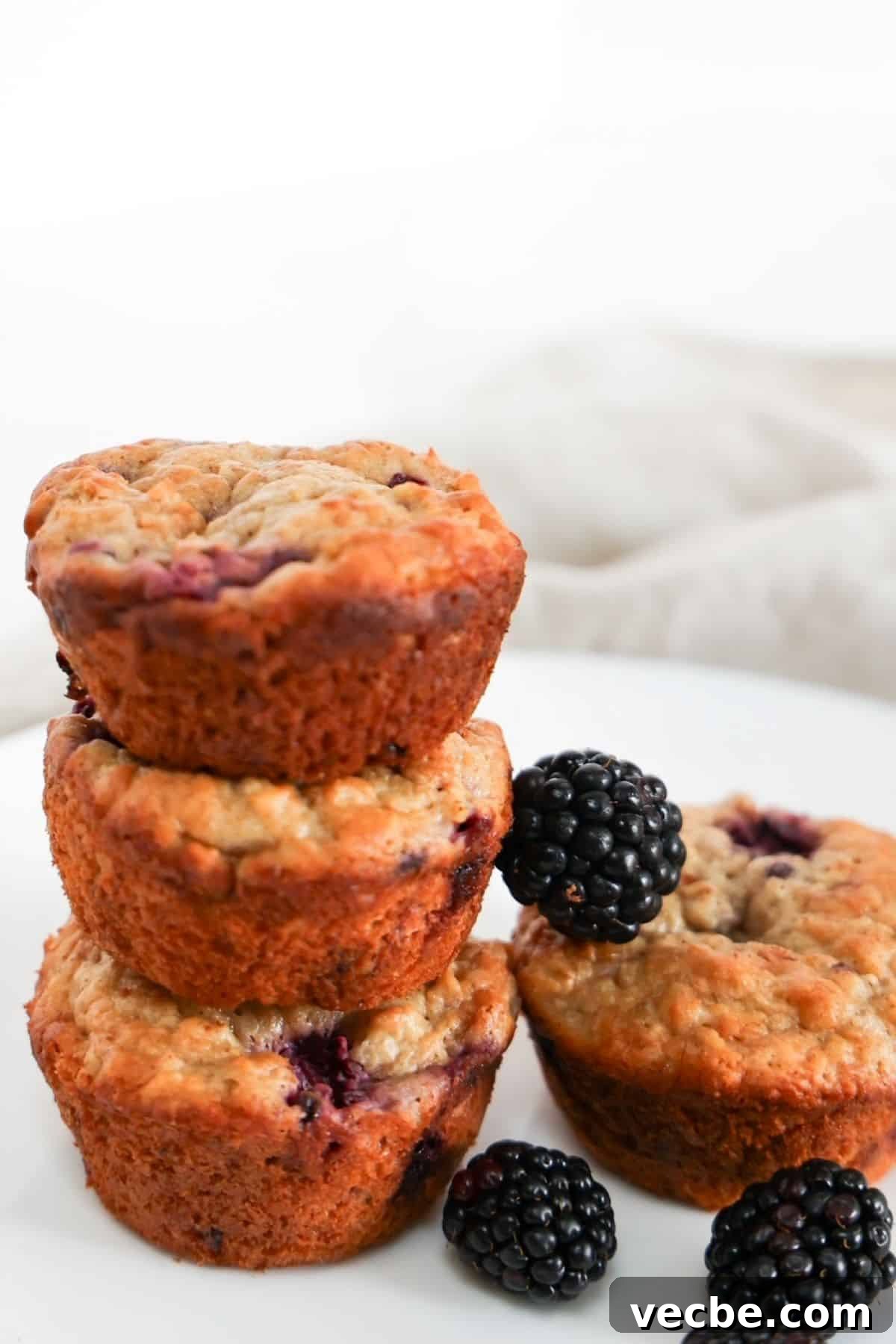 Banana Blackberry Oatmeal Muffins Recipe 5 Four Blackberry Banana Oatmeal Muffins, three are stacked to the left of one muffin. A few blackberries are scattered around.