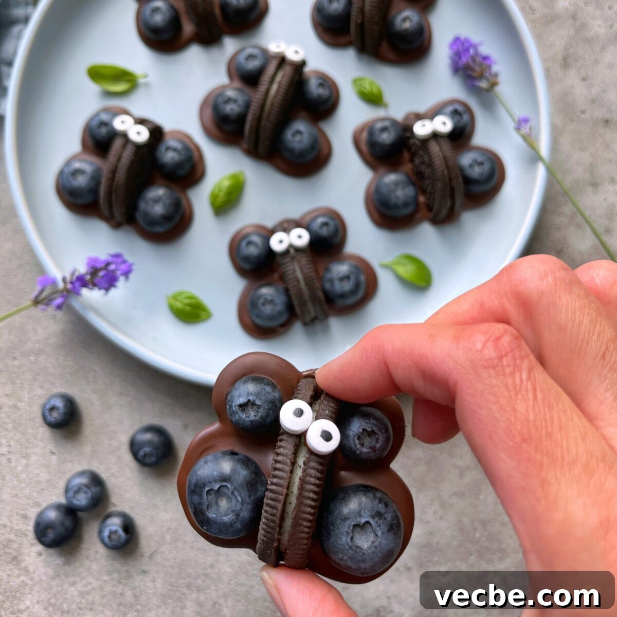 How to Make Chocolate Butterflies for Desserts 3 A hand holds a butterfly-shaped treat made with chocolate, blueberries, an Oreo cookie, and candy eyes. Similar bites rest on a plate in the background.
