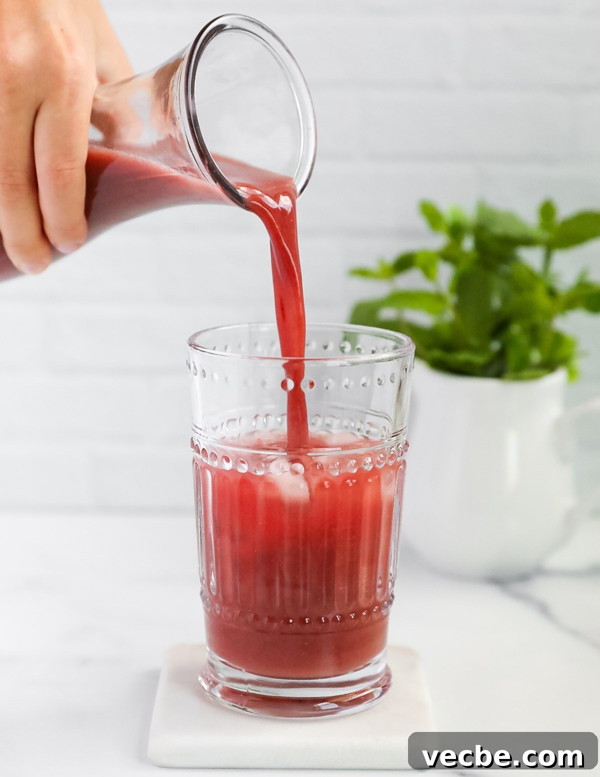 Strawberry Acai Lemonade Recipe 5 A hand pouring a pitcher of bright pink lemonade into a clear glass