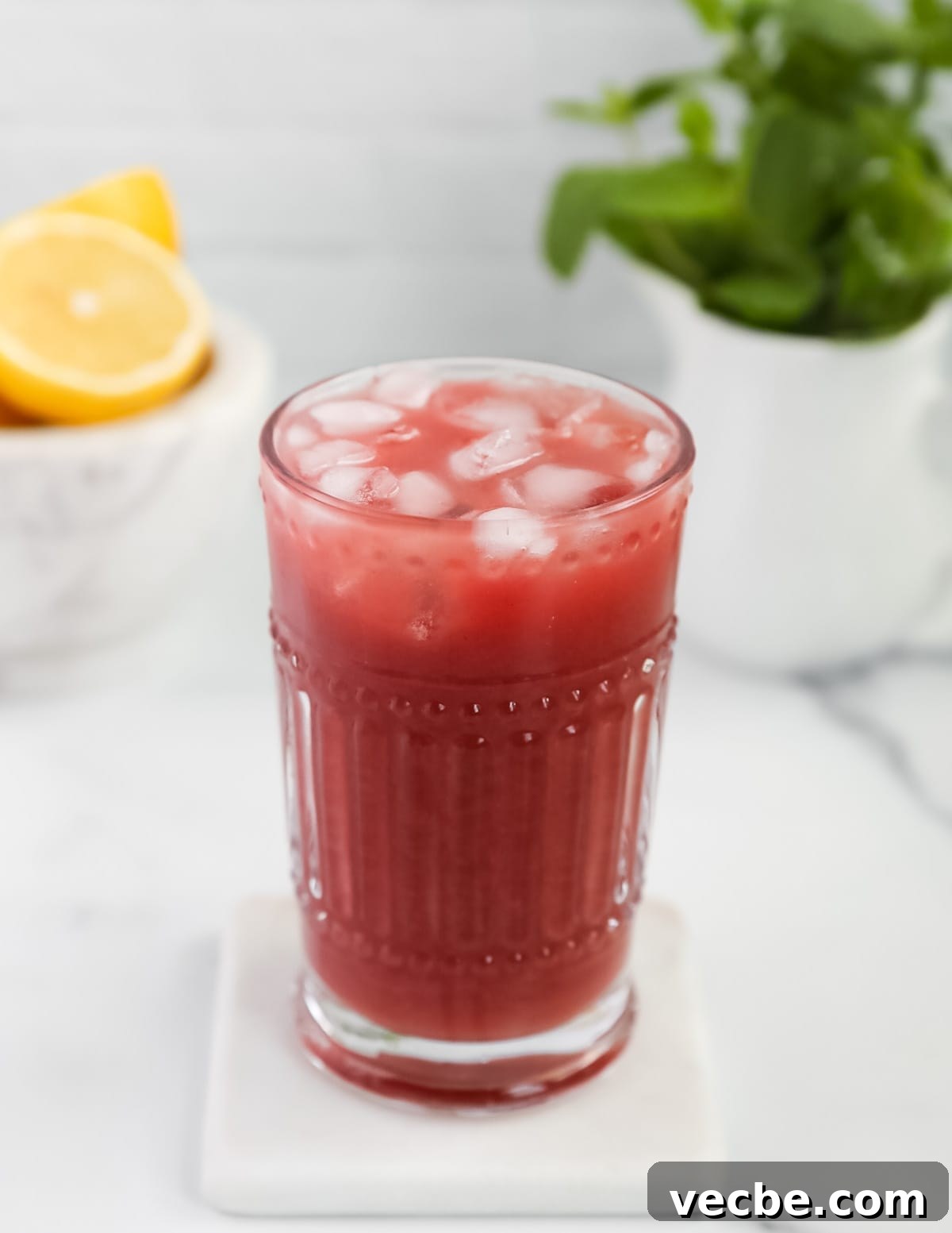 Strawberry Acai Lemonade Recipe 6 A picture of a clear glass filled with ice and pink lemonade. There is fresh mint and lemons in the background.