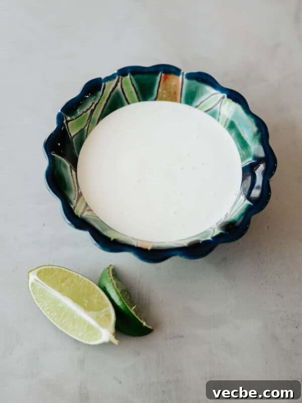 How to Make Mexican Chamoy Sauce at Home 11 blue and green painted scalloped bowl filled with homemade crema mexicana on a grey table with a few fresh lime wedges to the side.