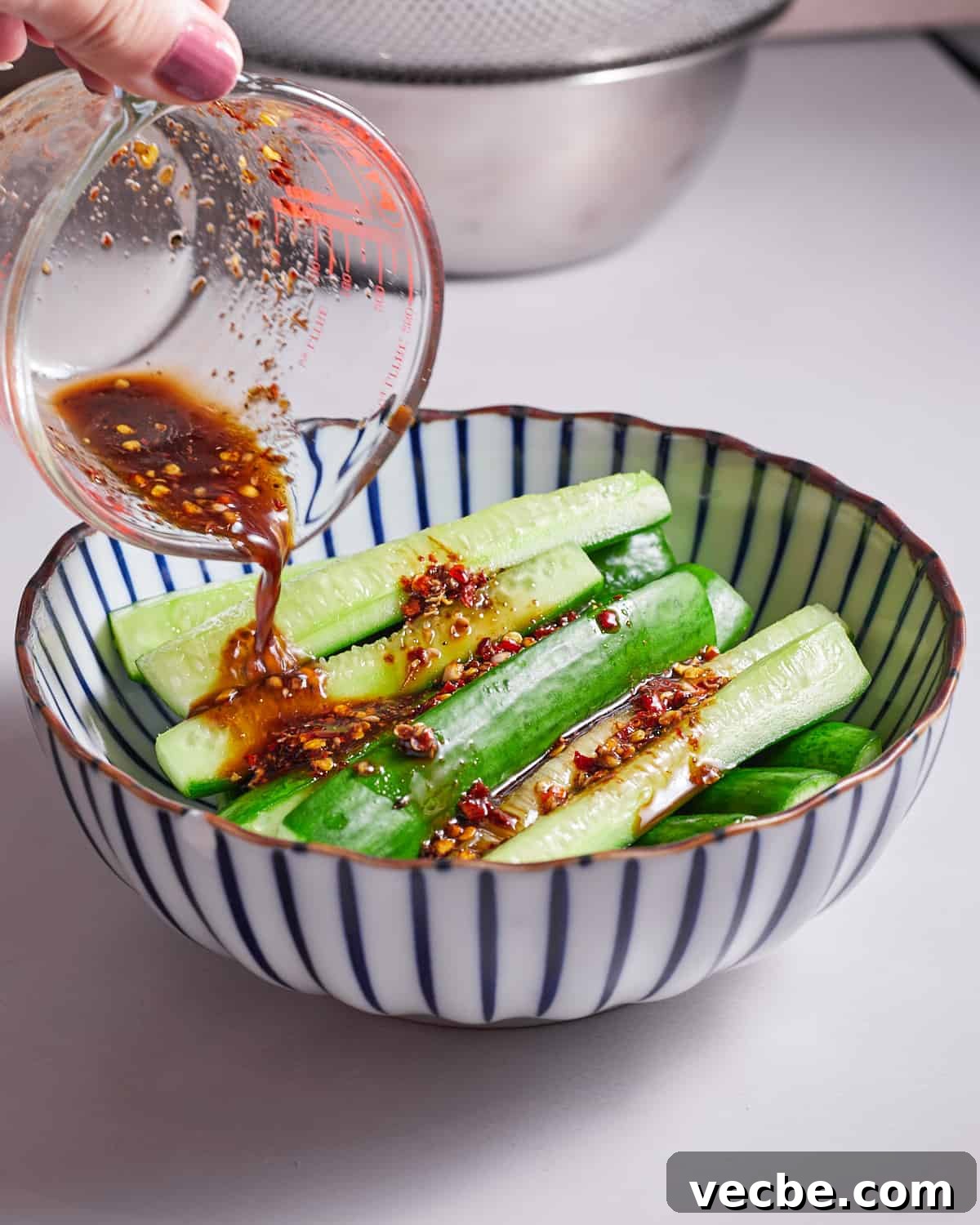 Quick Chinese Pickled Cucumbers Recipe 9 Photo of someone pouring the marinade over the cucumbers in a stripy white and blue bowl.