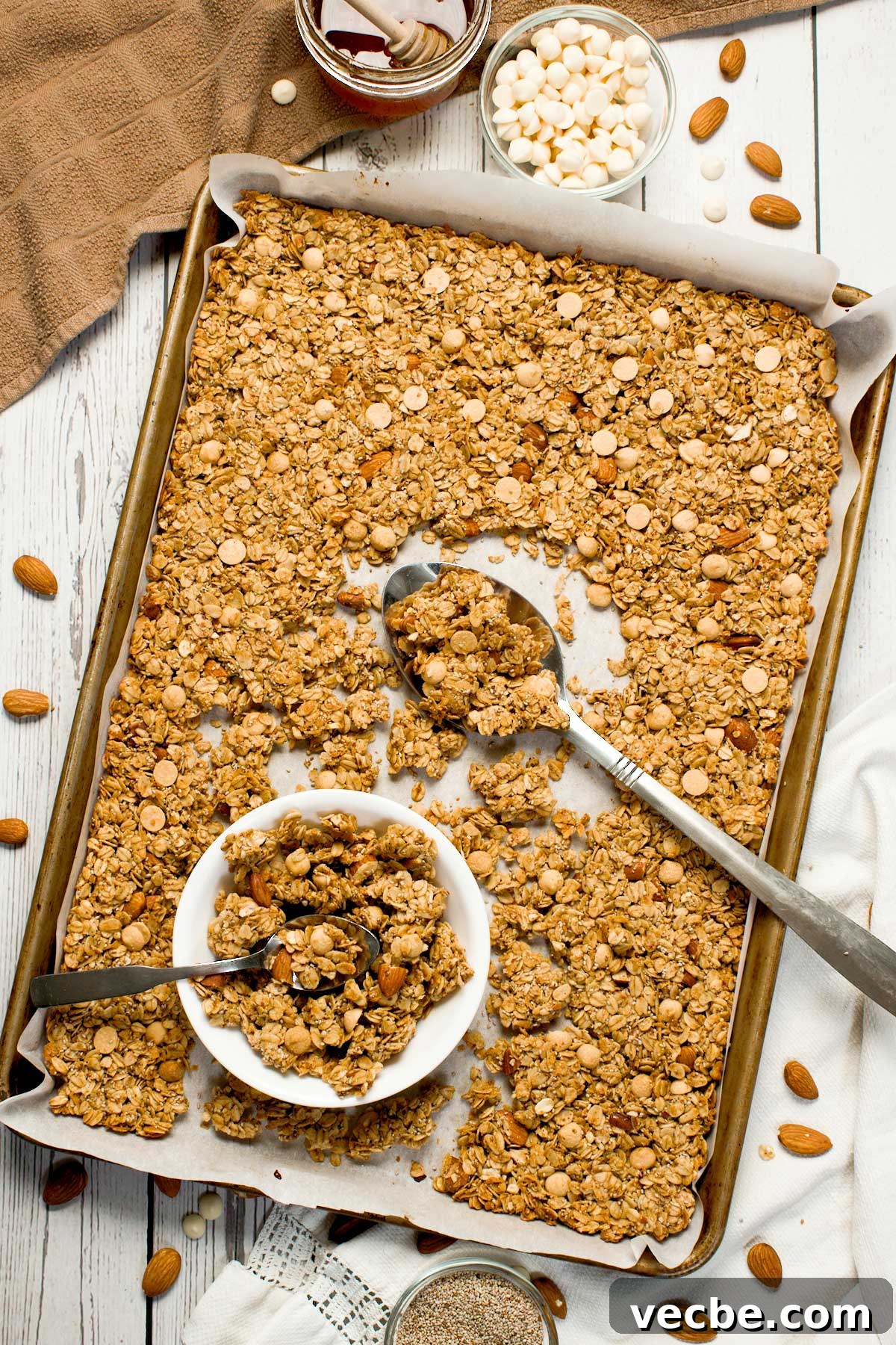 White Chocolate Vanilla Almond Granola 3 The top view of almond granola with white chocolate chips in a baking sheet with a large spoon and a small white bowl.