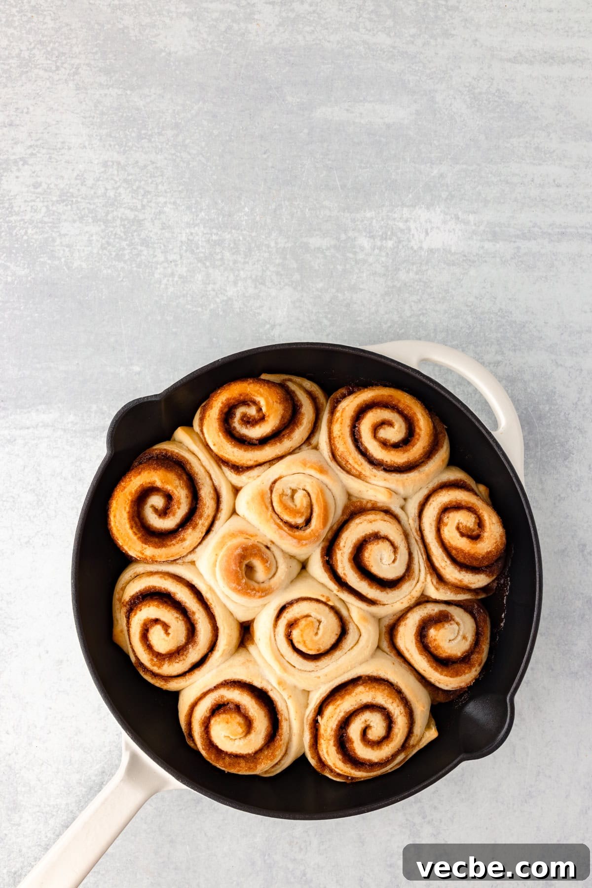 A top view of freshly baked cinnamon rolls arranged in a circular pattern in a round skillet. The rolls are golden brown and neatly spiraled, sitting on a light, textured surface.