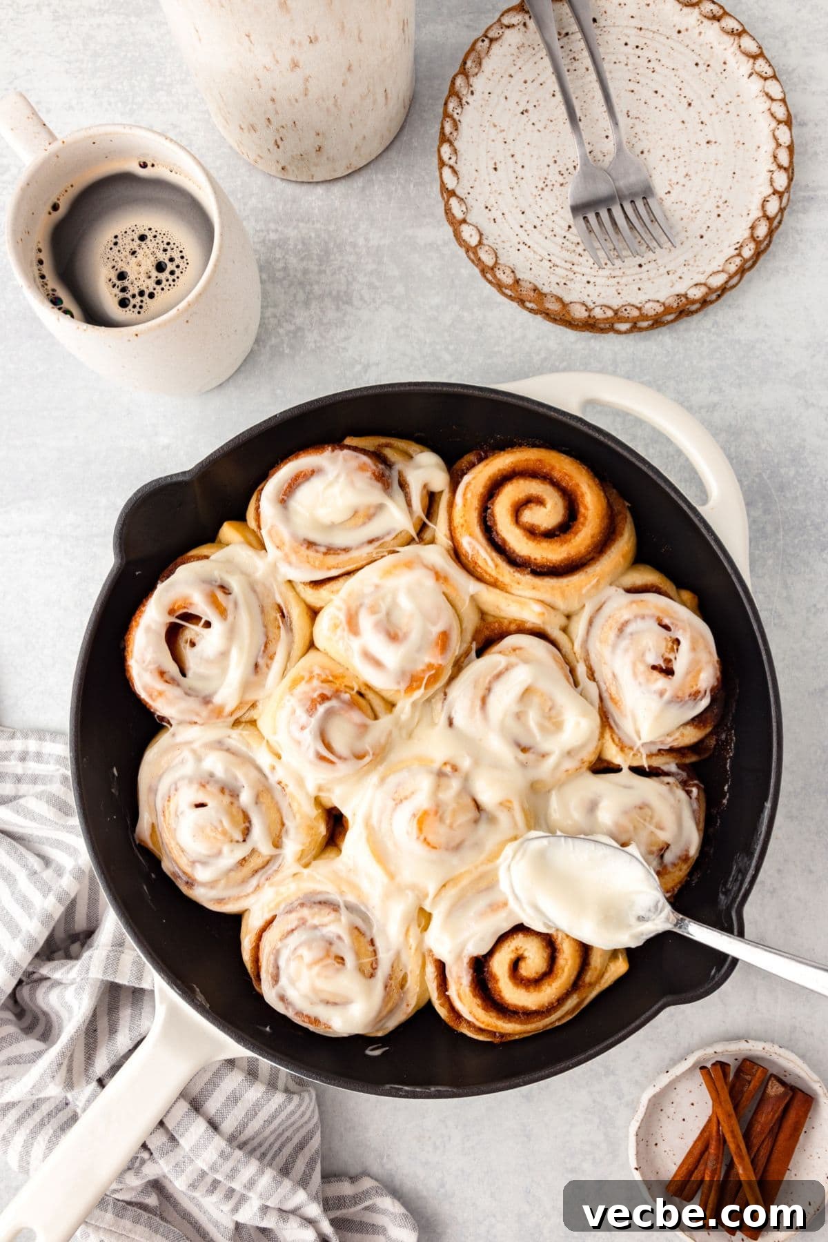 A pan of freshly baked cinnamon rolls topped with creamy icing, accompanied by a cup of coffee, a striped napkin, a small plate with two forks, and a dish with cinnamon sticks on a light surface.