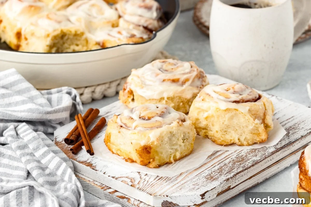 Freshly baked cinnamon rolls with white icing on a wooden board, accompanied by cinnamon sticks. A striped cloth and a cup of coffee are nearby, enhancing the cozy breakfast setting.