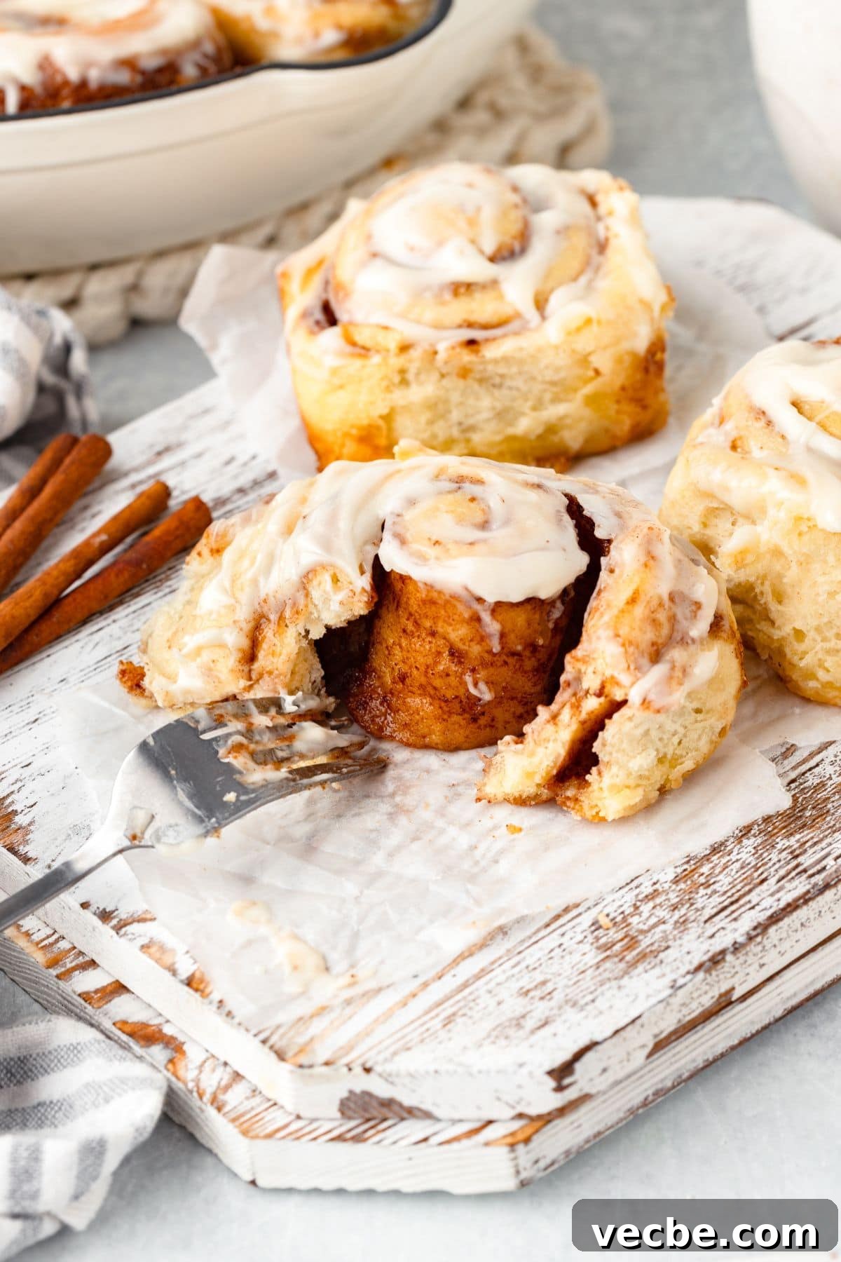 Close-up of cinnamon rolls with icing on a wooden board. One roll is partially cut with a metal spatula beneath it. Cinnamon sticks are beside the rolls, and a striped cloth is visible in the background.