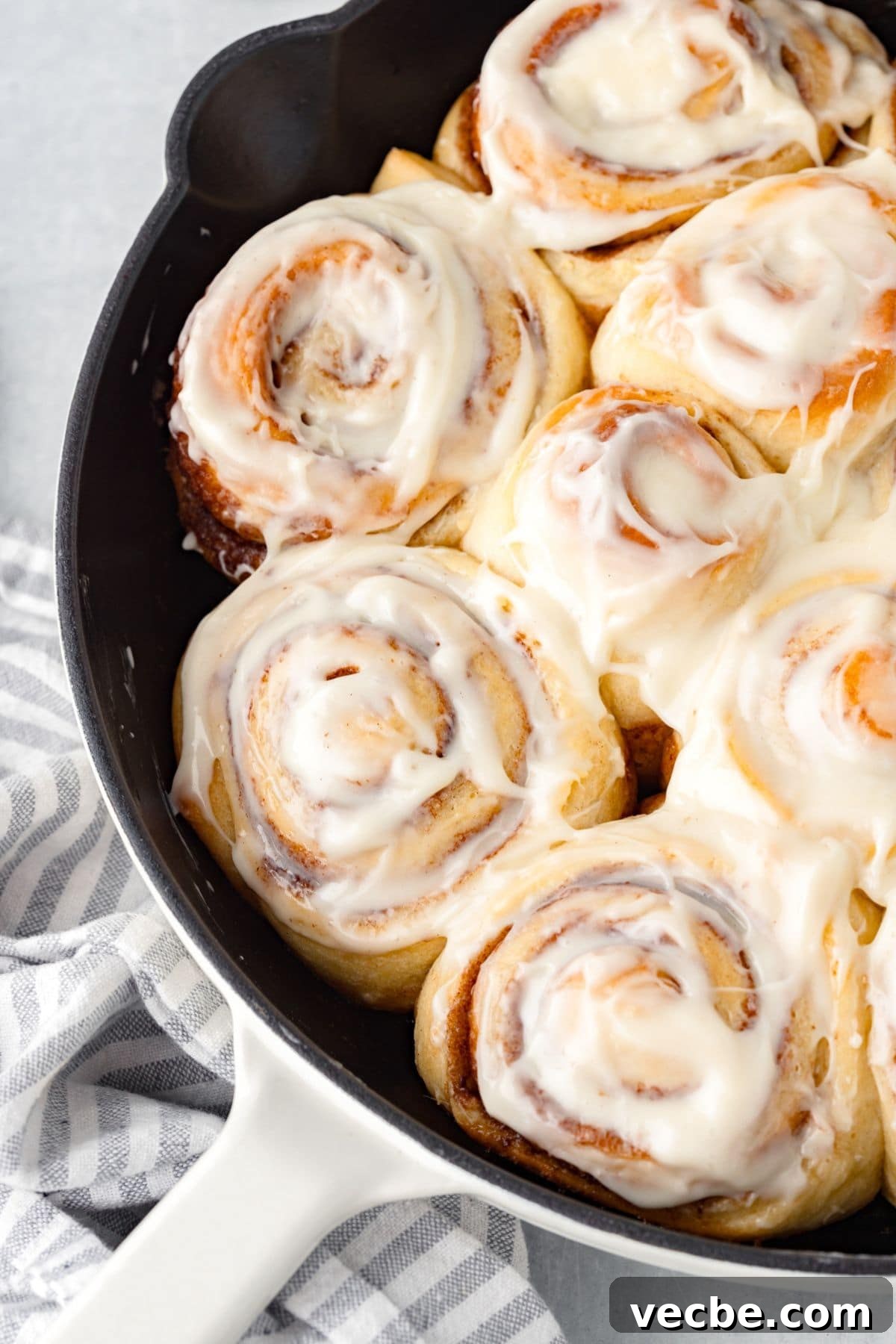 A baking pan filled with freshly baked cinnamon rolls topped with white icing. The rolls are golden brown and arranged closely together, offering a gooey and appetizing appearance. A striped cloth is partially visible underneath the pan.