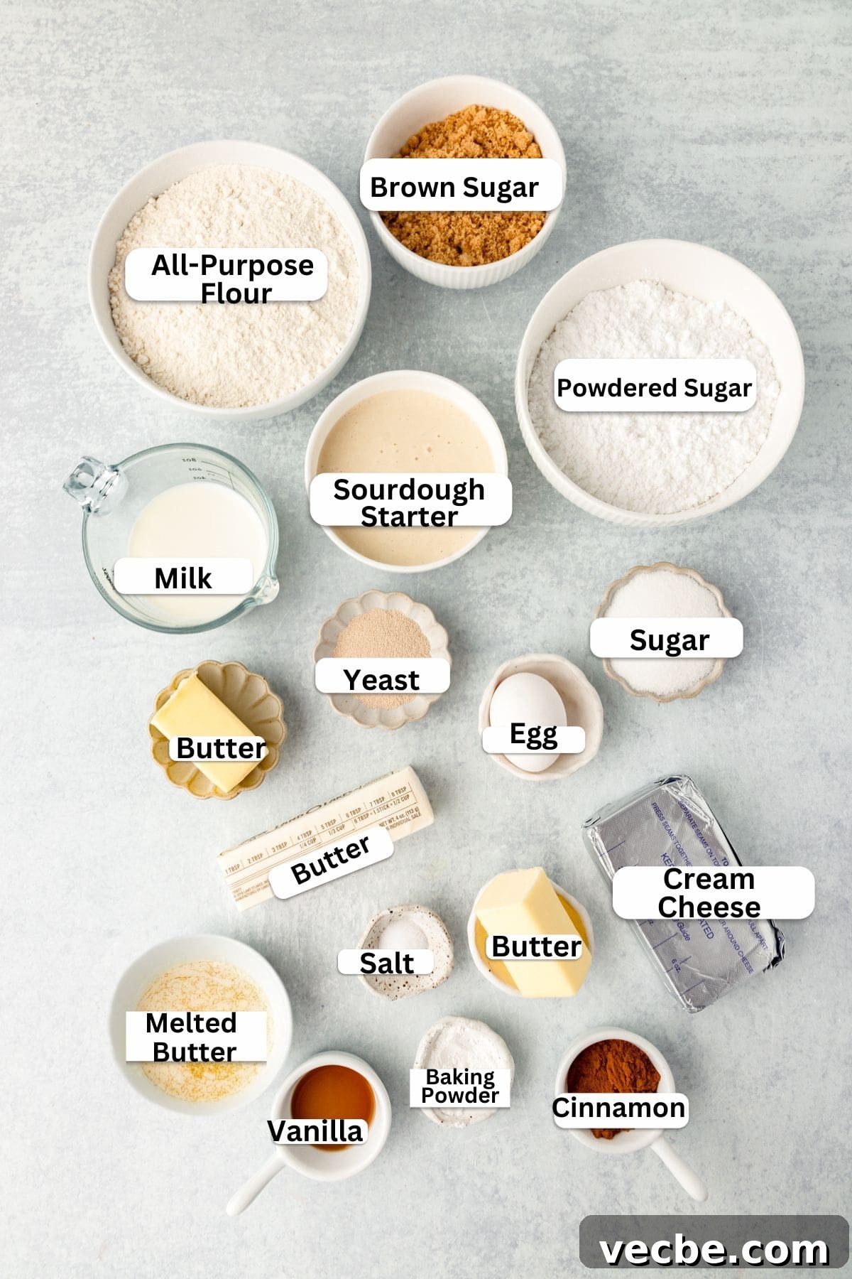 An assortment of baking ingredients labeled and arranged on a light surface, including bowls of flour, sugars, sourdough starter, milk, yeast, butter, egg, cream cheese, melted butter, salt, vanilla, baking powder, and cinnamon.
