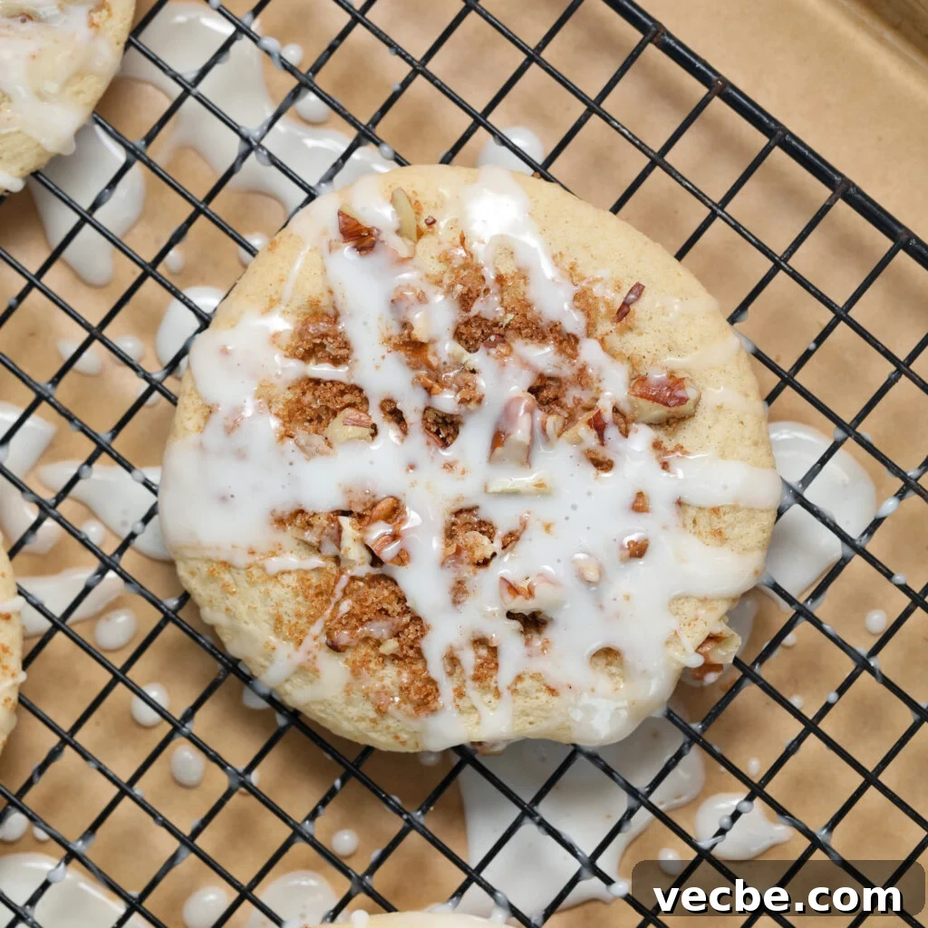 Coffee Cake Cookies with Cinnamon Streusel 2 A close-up of a round, baked coffee cake donut topped with crumbled cinnamon filling and drizzled with white icing, resting on a black wire cooling rack lined with brown parchment paper.