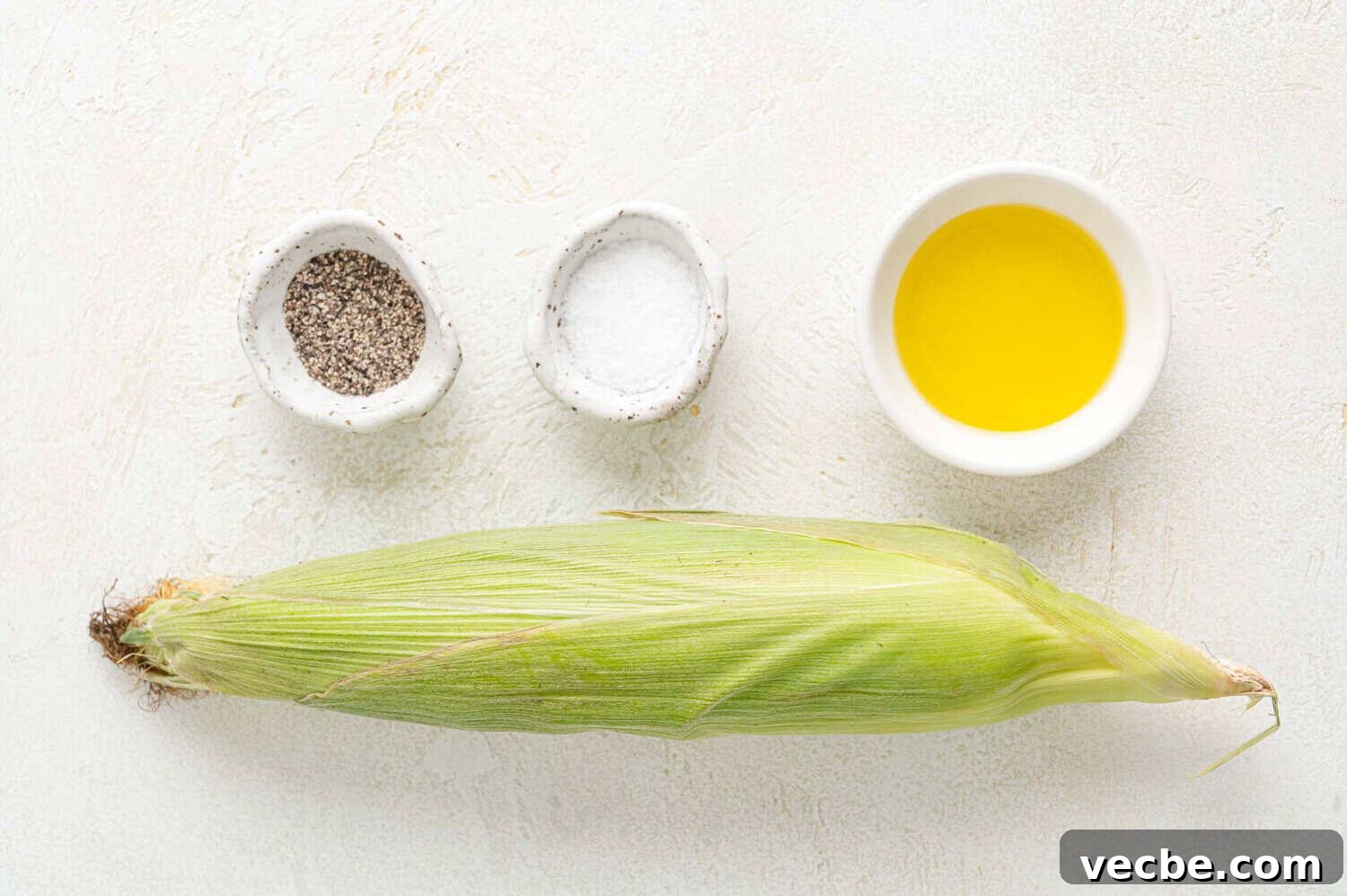 Crispy Buttered Air Fryer Corn on the Cob 2 An ear of corn in husk is placed on a white surface with small bowls of black pepper, salt, and olive oil arranged above it.