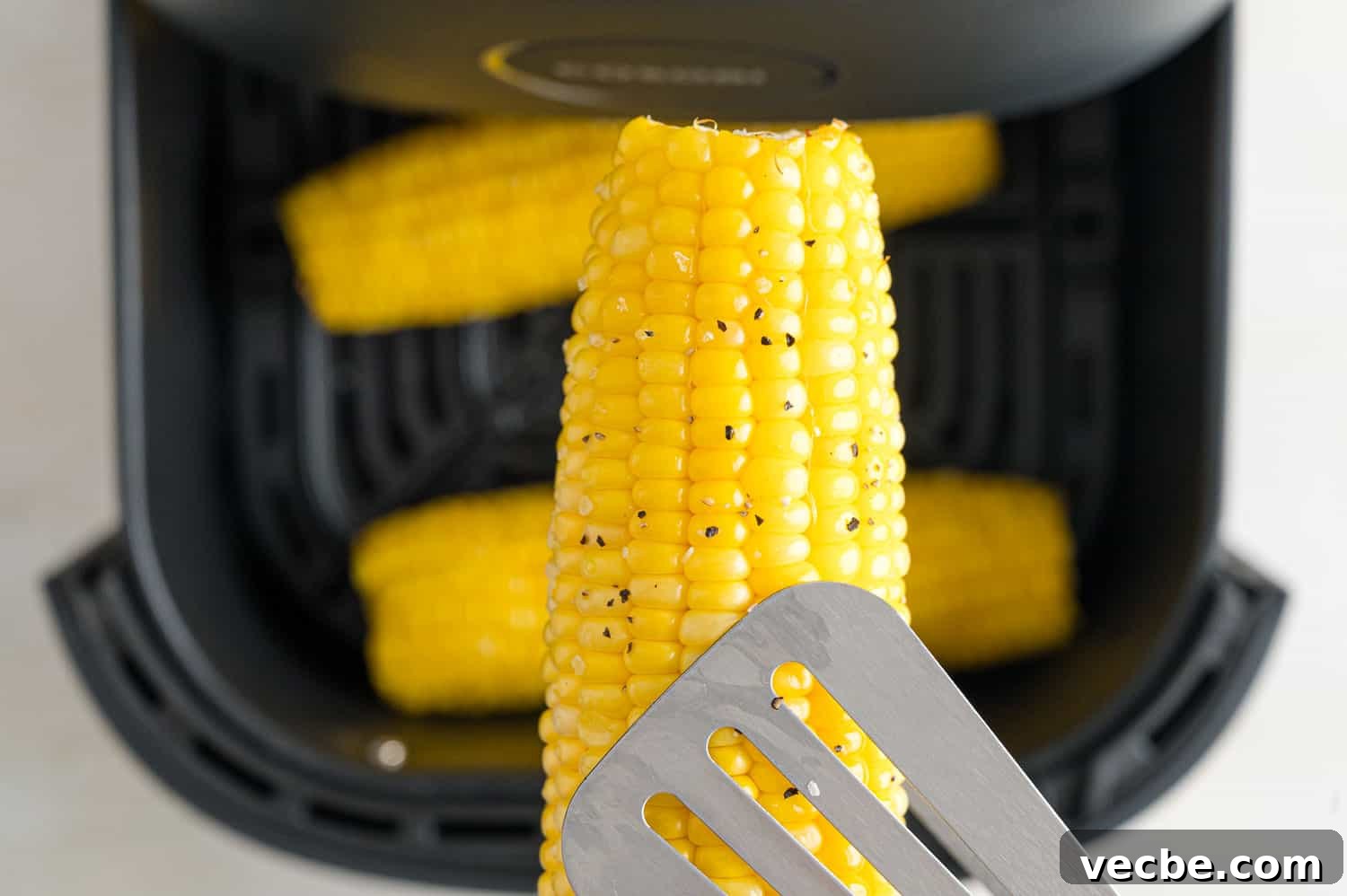 Crispy Buttered Air Fryer Corn on the Cob 7 Close up of tongs holding an ear of corn above more corn inside the air fryer.