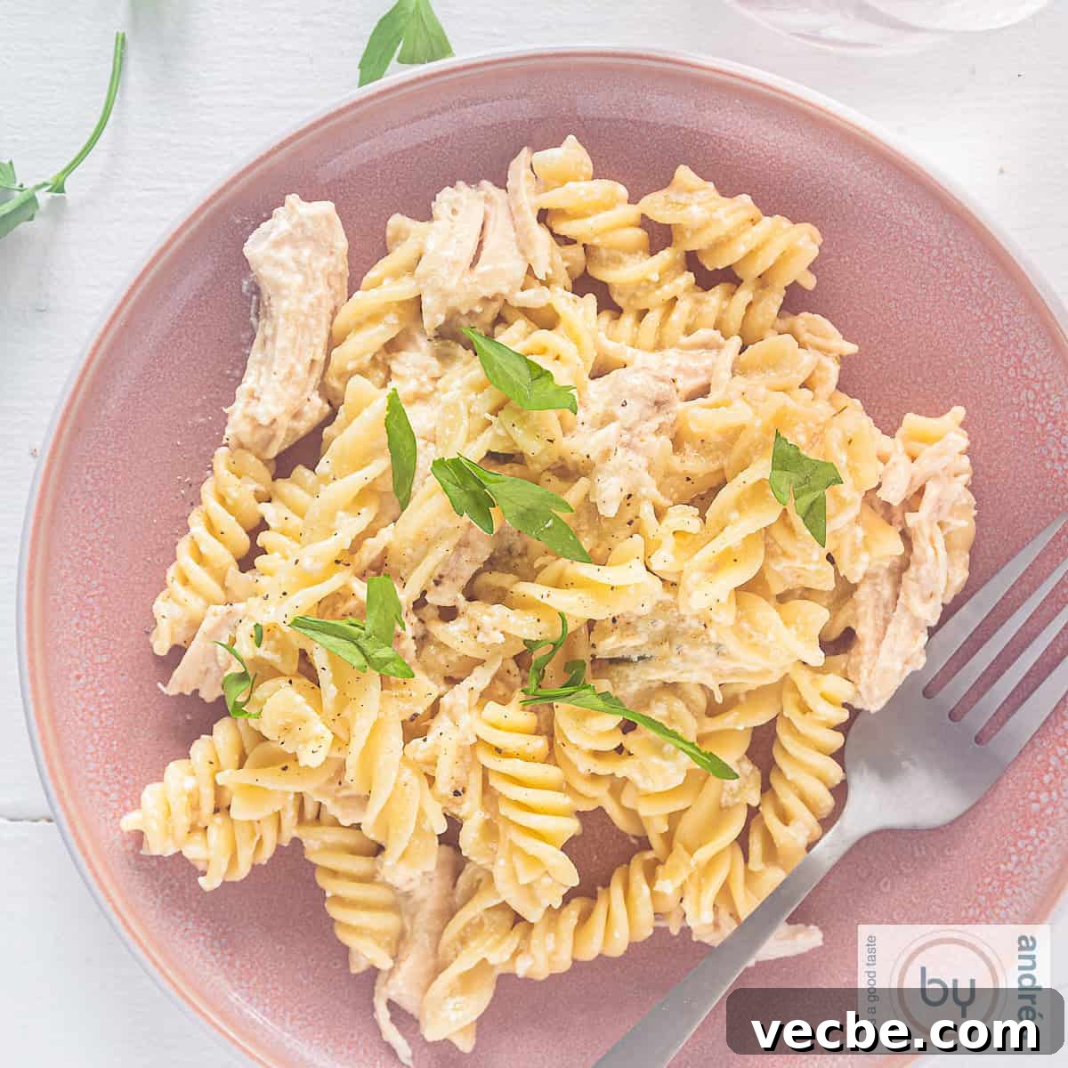 Slow Cooker Garlic Parmesan Chicken with Fusilli 6 A square photo of a pink plate with pasta, creamy sauce with Parmesan and garlic and parsley on a white background